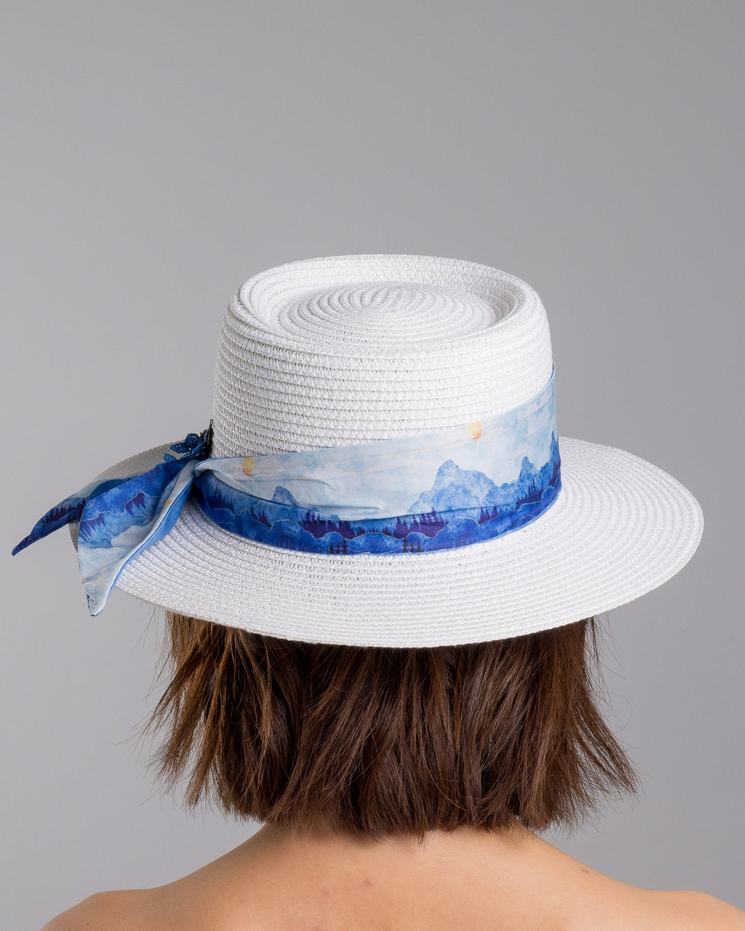 The model is showcasing the back side close up of the white woven straw boater hat with a wide brim, wrapped in a soft blue watercolour scarf and finished with delicate blue beaded floral embellishments at the side. Designed by Merve Bayindir millinery.