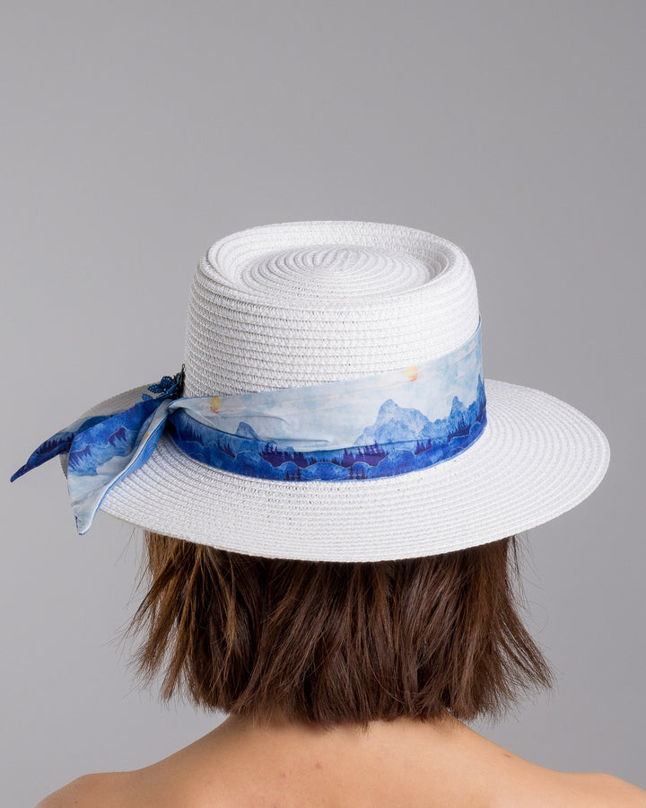 The model is showcasing the back side close up of the white woven straw boater hat with a wide brim, wrapped in a soft blue watercolour scarf and finished with delicate blue beaded floral embellishments at the side. Designed by Merve Bayindir millinery.