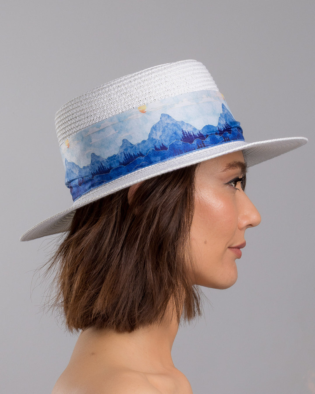 The model is showcasing the right side close up of the white woven straw boater hat with a wide brim, wrapped in a soft blue watercolour scarf and finished with delicate blue beaded floral embellishments at the side. Designed by Merve Bayindir millinery.