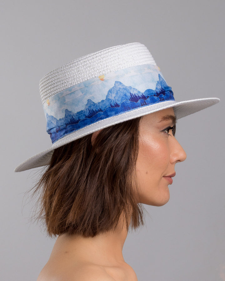 The model is showcasing the right side close up of the white woven straw boater hat with a wide brim, wrapped in a soft blue watercolour scarf and finished with delicate blue beaded floral embellishments at the side. Designed by Merve Bayindir millinery.