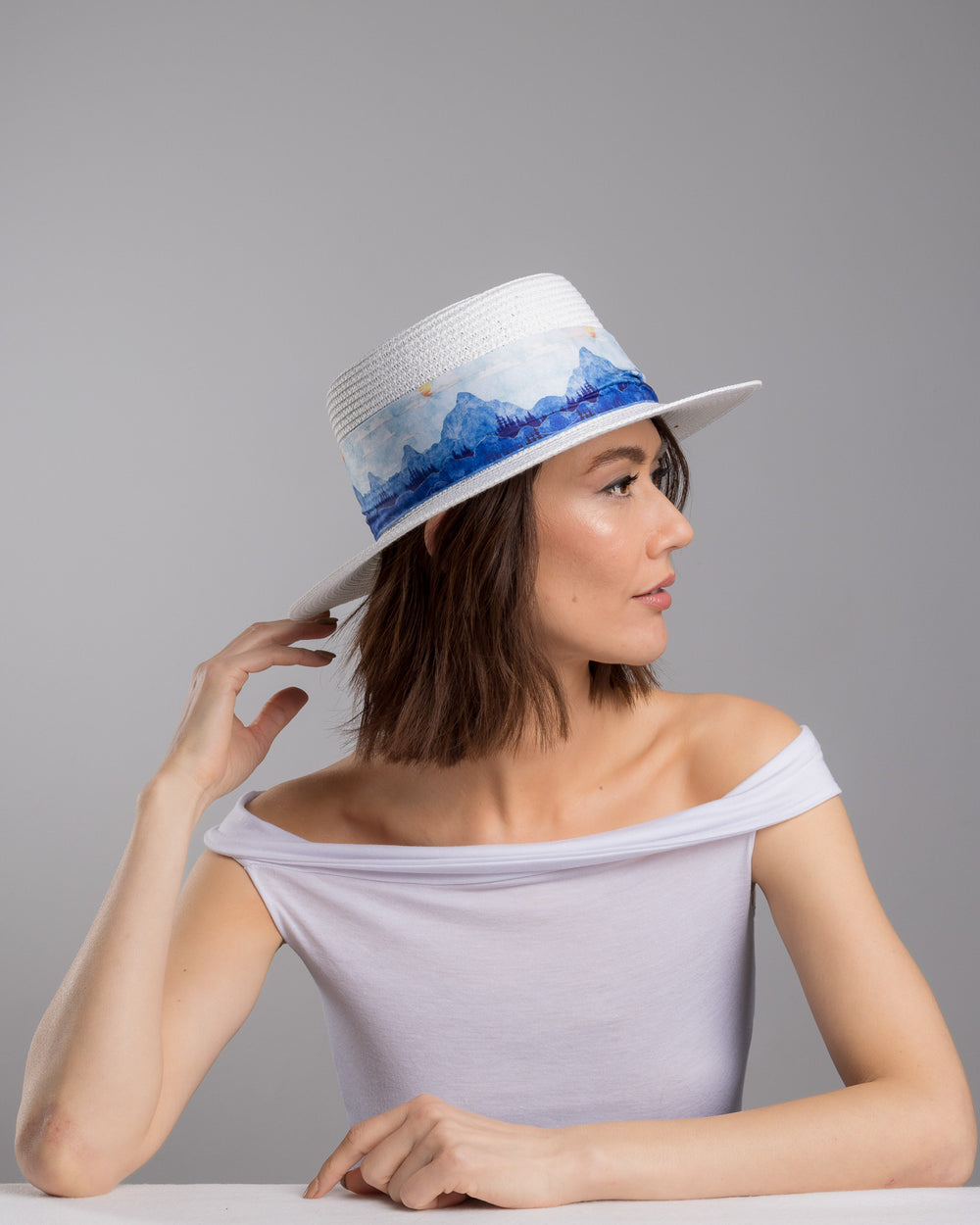 The female model is posing, wearing , Azuril,  the white woven straw boater hat with a wide brim, wrapped in a soft blue watercolour scarf and finished with delicate blue beaded floral embellishments at the side. Designed by Merve Bayindir millinery.