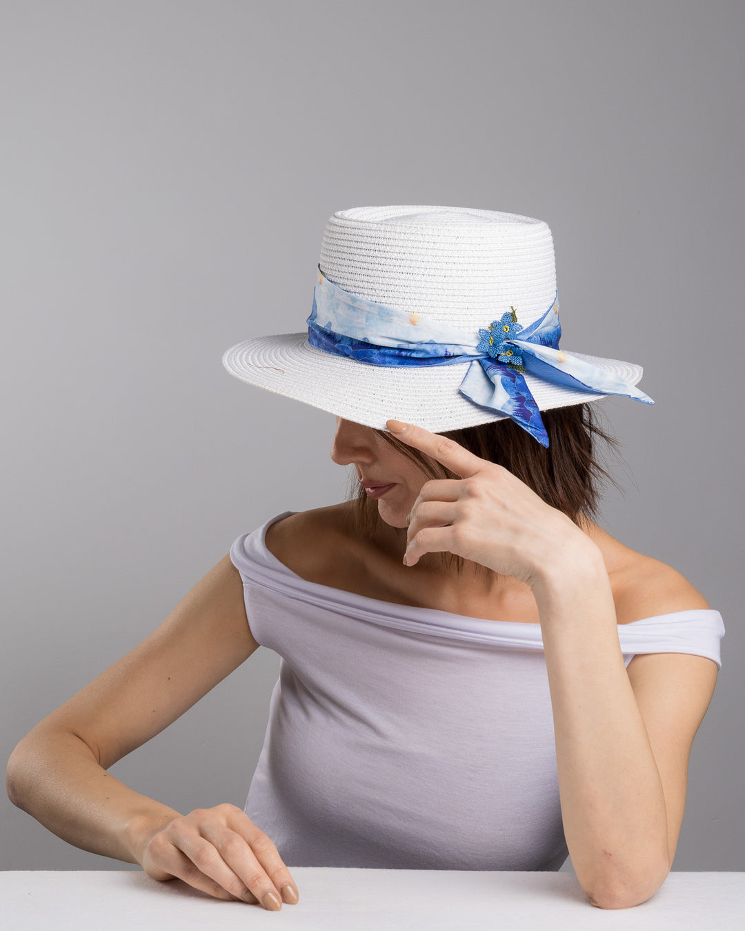 The female model is posing, wearing , Azuril,  the white woven straw boater hat with a wide brim, wrapped in a soft blue watercolour scarf and finished with delicate blue beaded floral embellishments at the side. Designed by Merve Bayindir millinery.