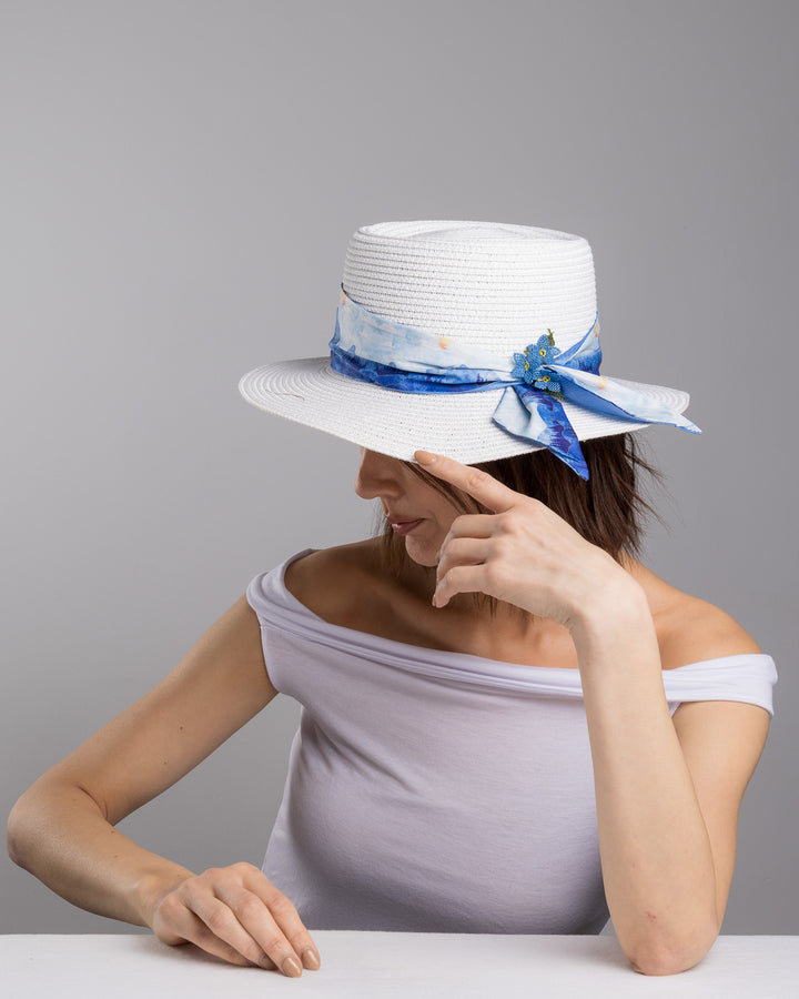 The female model is posing, wearing , Azuril,  the white woven straw boater hat with a wide brim, wrapped in a soft blue watercolour scarf and finished with delicate blue beaded floral embellishments at the side. Designed by Merve Bayindir millinery.