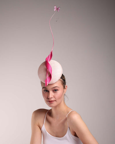 Model wearing a light pink button shape fascinator centred to her head. The fascinator is decorated with hot pink swirl integrated with a pink swirl of ostrich quill in the center. At the tip of the quill swirl there is a pink organza butterfly.