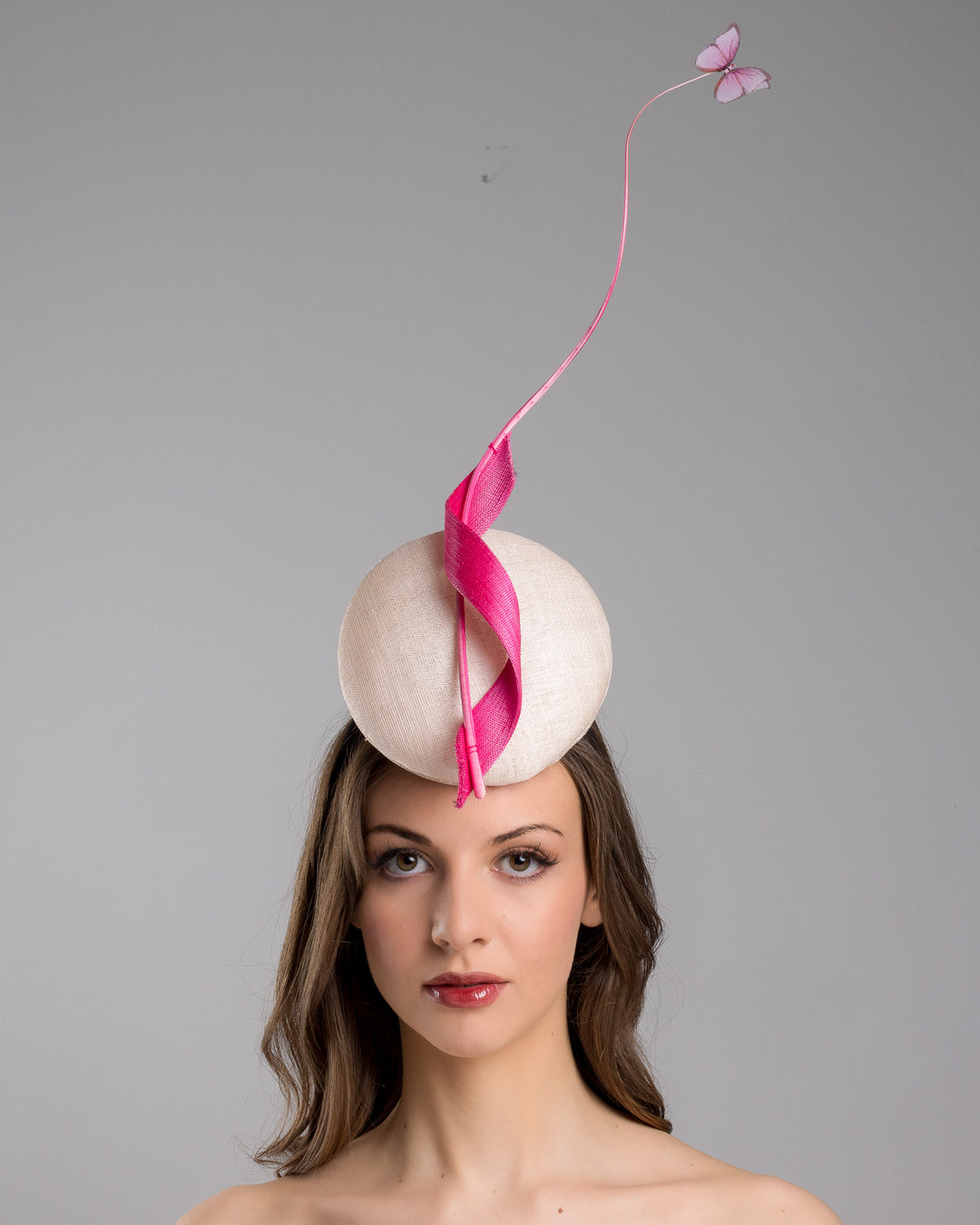 Model is showcasing the front side of the light pink button shape fascinator attached to a headband. The fascinator is decorated with hot pink jinsin swirl integrated with a pink swirl of ostrich quill in the center. At the tip of the quill swirl there is a pink organza butterfly.