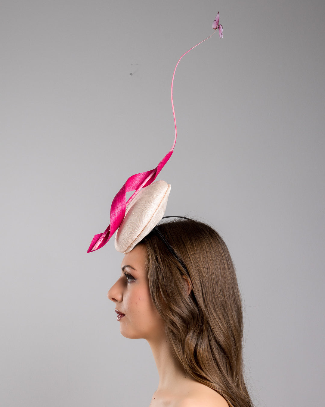 Model is showcasing the left side of the light pink button shape fascinator attached to a headband. The fascinator is decorated with hot pink jinsin swirl integrated with a pink swirl of ostrich quill in the center. At the tip of the quill swirl there is a pink organza butterfly.