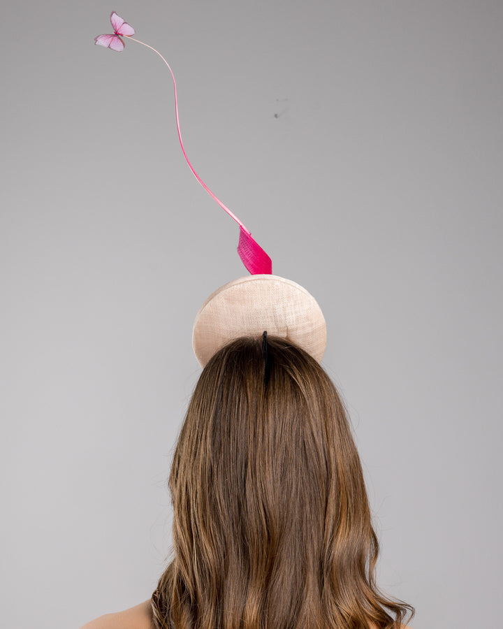 Model is showcasing the back of the light pink button shape fascinator attached to a headband. The fascinator is decorated with hot pink jinsin swirl integrated with a pink swirl of ostrich quill in the center. At the tip of the quill swirl there is a pink organza butterfly.