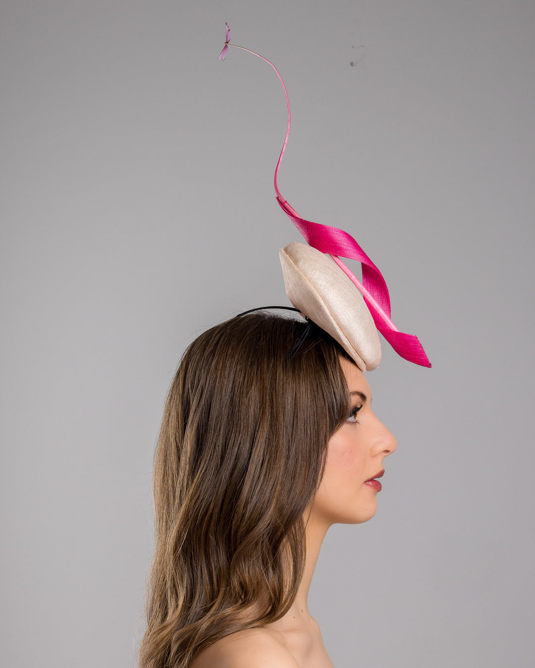 Model is showcasing the right side of the light pink button shape fascinator attached to a headband. The fascinator is decorated with hot pink jinsin swirl integrated with a pink swirl of ostrich quill in the center. At the tip of the quill swirl there is a pink organza butterfly.