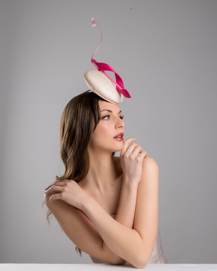 Model is posing wearing Merve Bayindir  light pink button shape fascinator attached to a headband. The fascinator is decorated with hot pink jinsin swirl integrated with a pink swirl of ostrich quill in the center. At the tip of the quill swirl there is a pink organza butterfly.