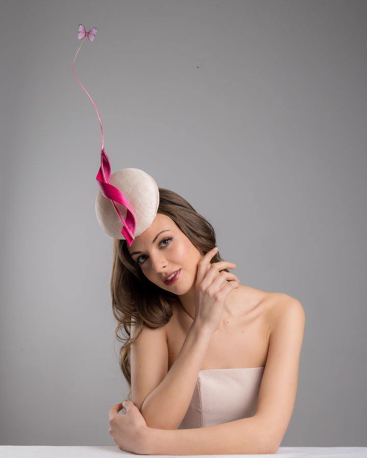 Model is posing wearing Merve Bayindir  light pink button shape fascinator attached to a headband. The fascinator is decorated with hot pink jinsin swirl integrated with a pink swirl of ostrich quill in the center. At the tip of the quill swirl there is a pink organza butterfly.