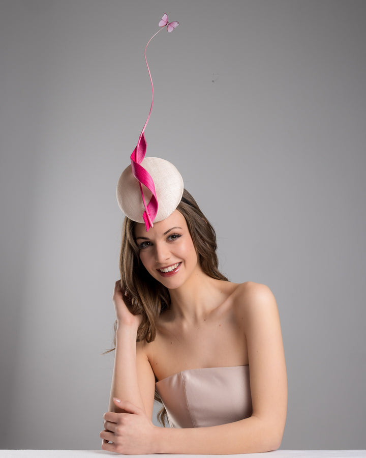 Model is posing wearing Merve Bayindir  light pink button shape fascinator attached to a headband. The fascinator is decorated with hot pink jinsin swirl integrated with a pink swirl of ostrich quill in the center. At the tip of the quill swirl there is a pink organza butterfly.