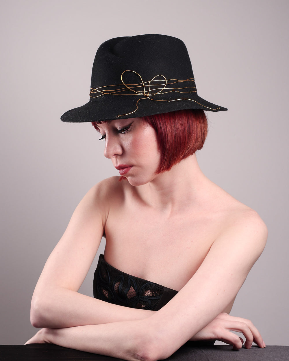 The model showcases th left side of the Ahether Fedora hat, a classic silhouette, crafted in black felt and adorned with freeform golden wirework, finished with an abstract metallic bow motif. Designed by Merve Bayindir.