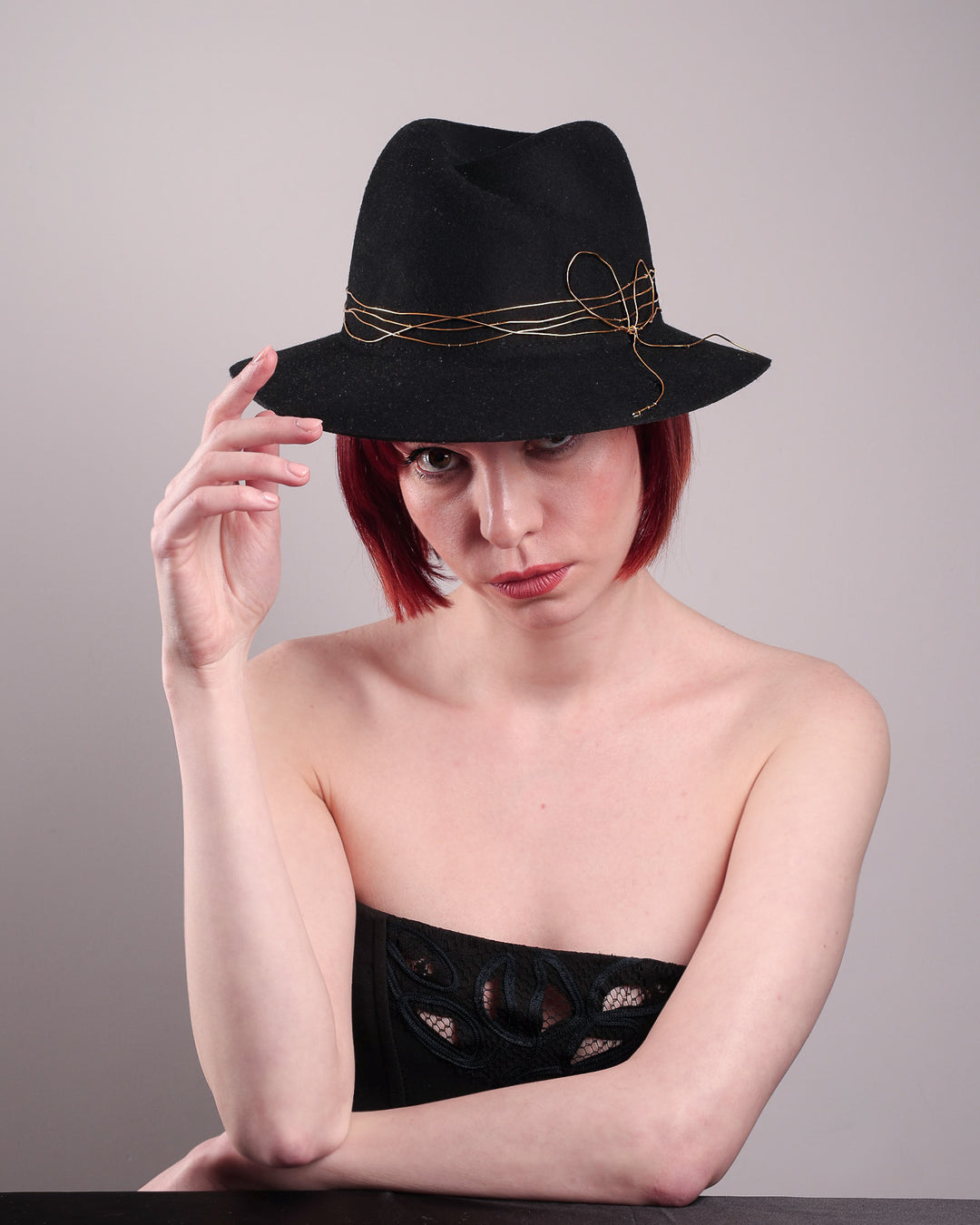 The model showcases the front of the Ahether Fedora hat, a classic silhouette, crafted in black felt and adorned with freeform golden wirework, finished with an abstract metallic bow motif. Designed by Merve Bayindir.