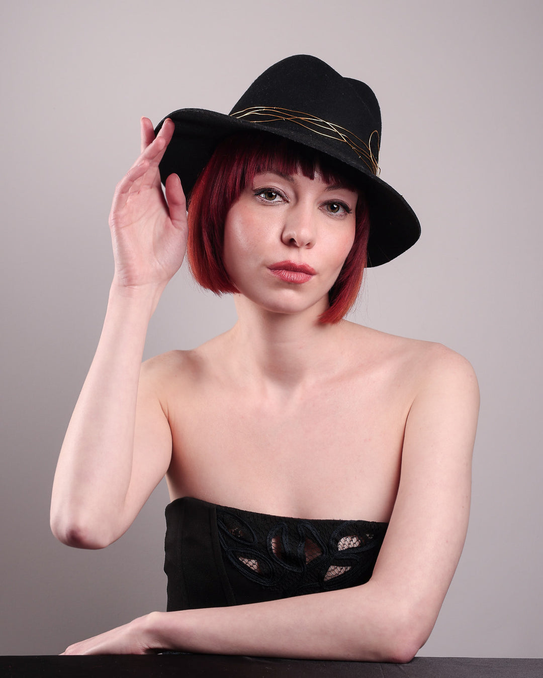 The model showcases the low angle front of the Ahether Fedora hat, a classic silhouette, crafted in black felt and adorned with freeform golden wirework, finished with an abstract metallic bow motif. Designed by Merve Bayindir.