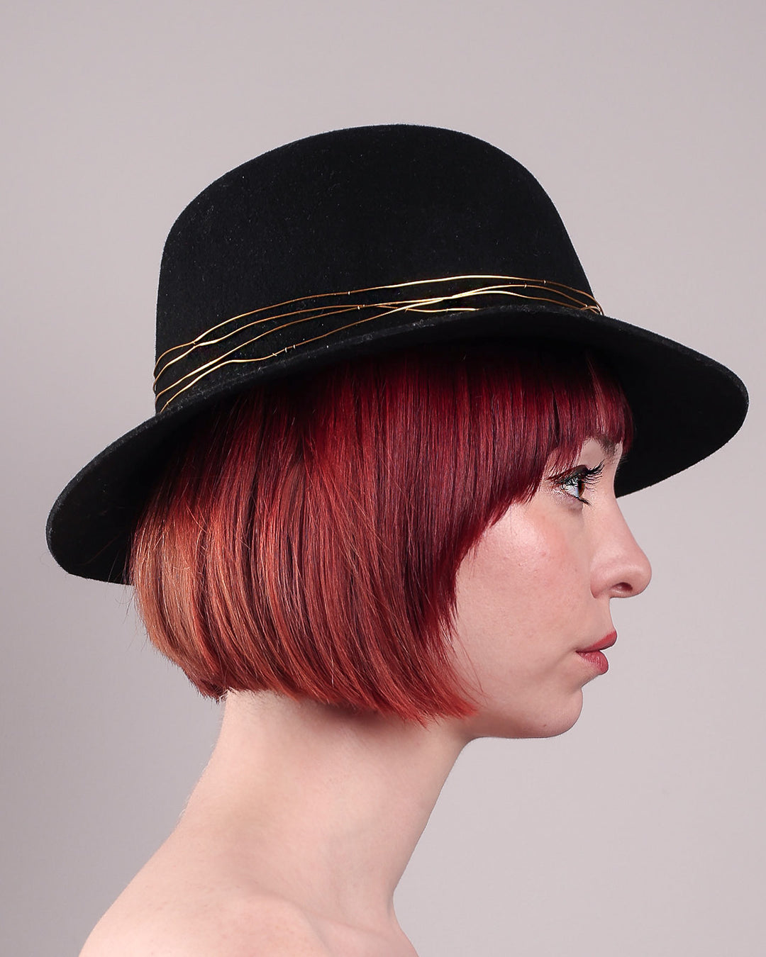 The model showcases the right side close up of the Ahether Fedora hat, a classic silhouette, crafted in black felt and adorned with freeform golden wirework, finished with an abstract metallic bow motif. Designed by Merve Bayindir.