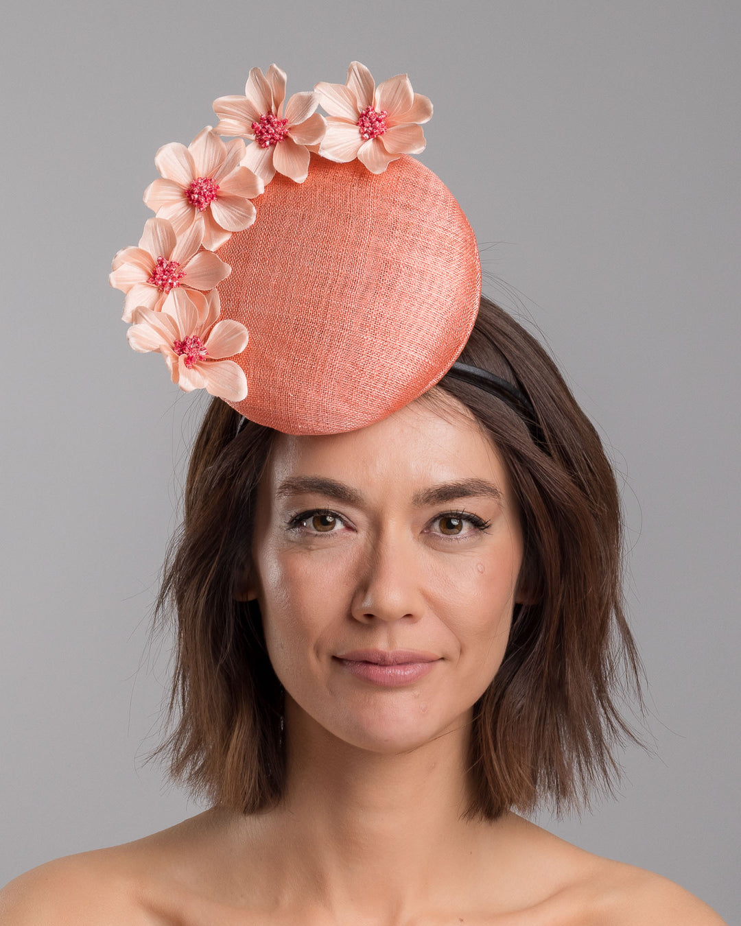 Model is showcasing the front side close up of coral coloured tall button shaped fascinator attached towards the right side of a headband. The fascinator is decorated from right side to the top with lighter coral coloured five flowers. Designed and made by Merve Bayindir millinery.