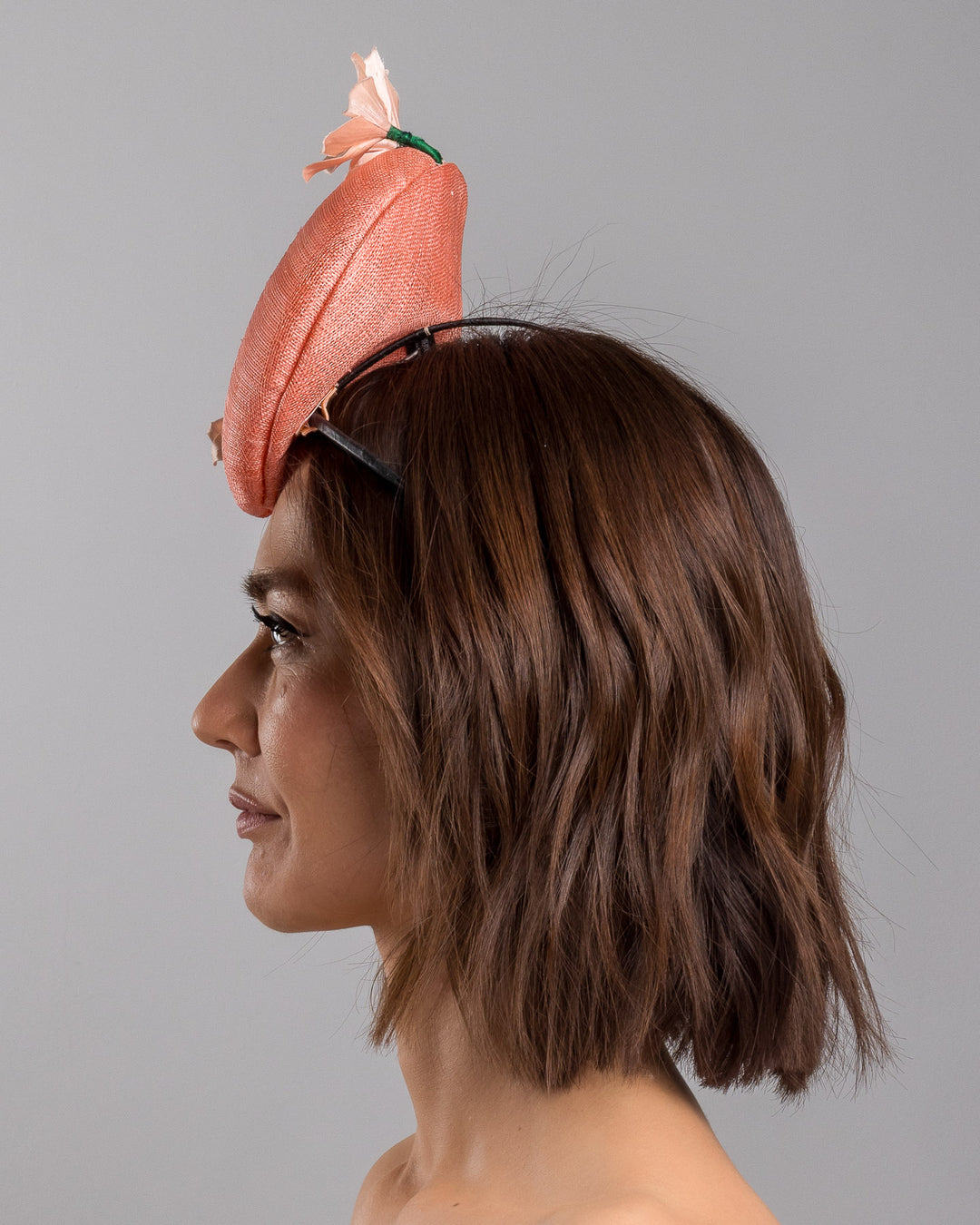 Model is showcasing the left side close up of coral coloured tall button shaped fascinator attached towards the right side of a headband. The fascinator is decorated from right side to the top with lighter coral coloured five flowers. Designed and made by Merve Bayindir millinery.