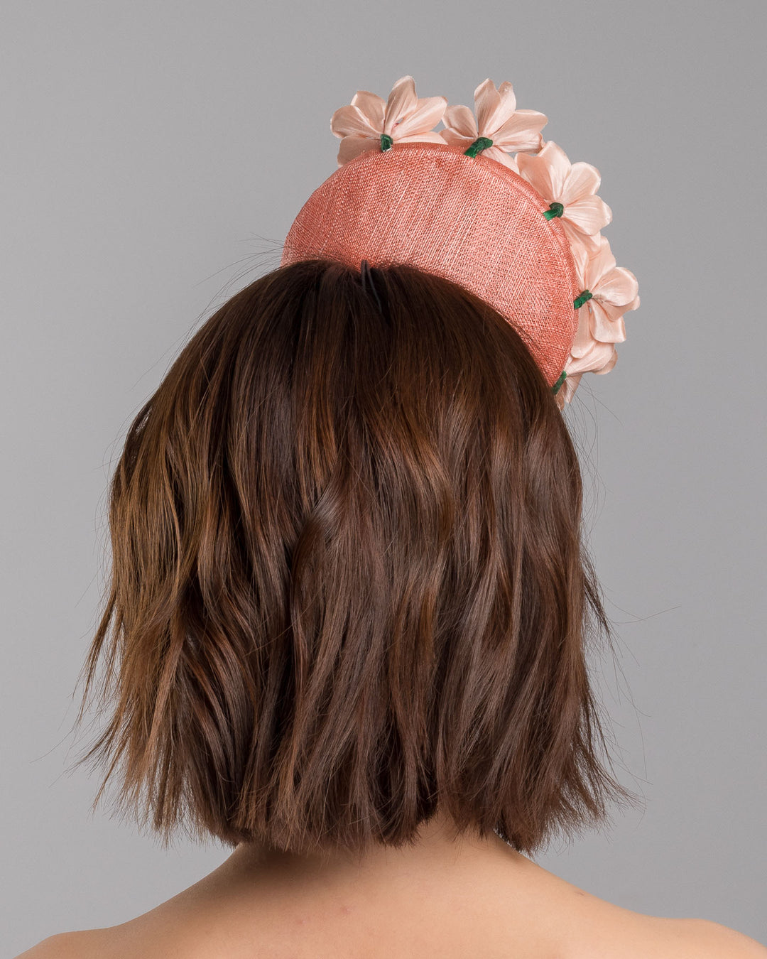 Model is showcasing the back side close up of coral coloured tall button shaped fascinator attached towards the right side of a headband. The fascinator is decorated from right side to the top with lighter coral coloured five flowers. Designed and made by Merve Bayindir millinery.