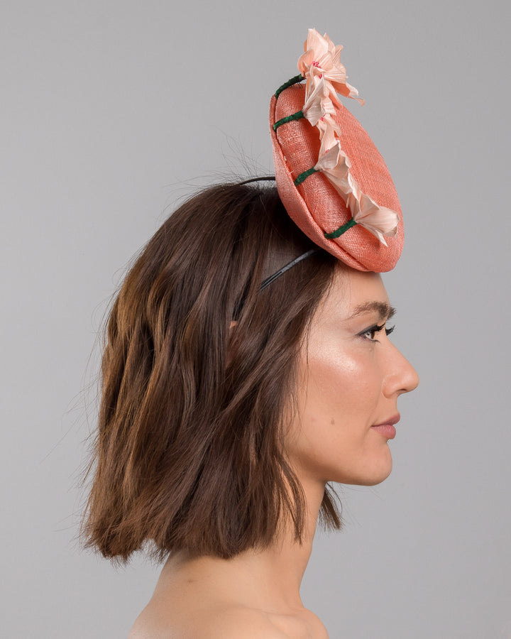 Model is showcasing the right side close up of coral coloured tall button shaped fascinator attached towards the right side of a headband. The fascinator is decorated from right side to the top with lighter coral coloured five flowers. Designed and made by Merve Bayindir millinery.
