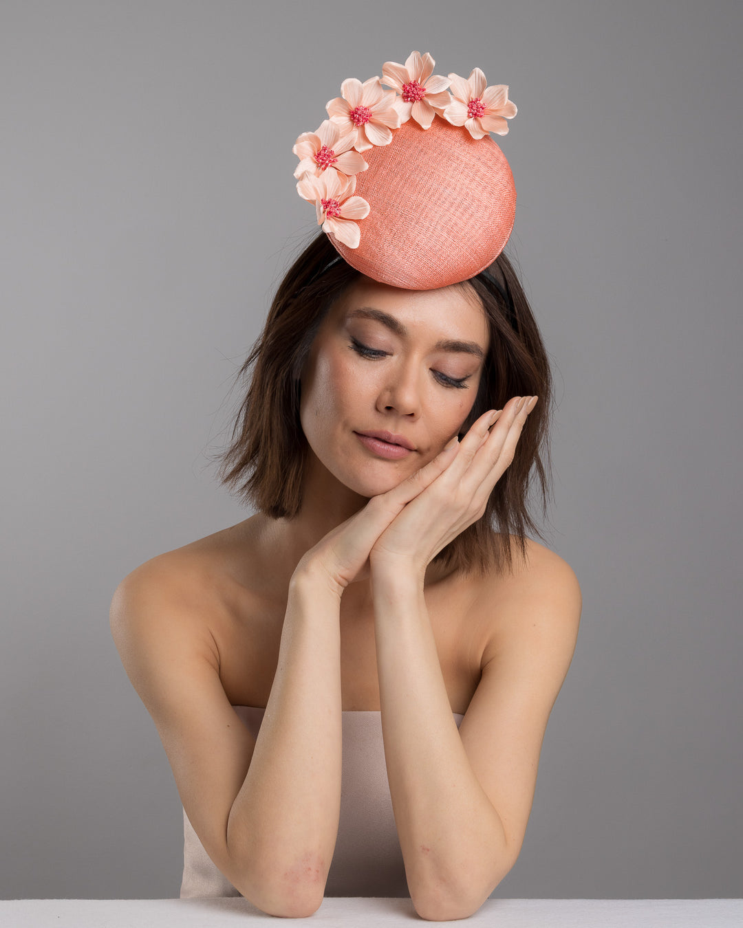 Female model is posing wearing Ainsley coral coloured tall button shaped fascinator attached towards the right side of a headband. The fascinator is decorated from right side to the top with lighter coral coloured five flowers. Designed and made by Merve Bayindir millinery.