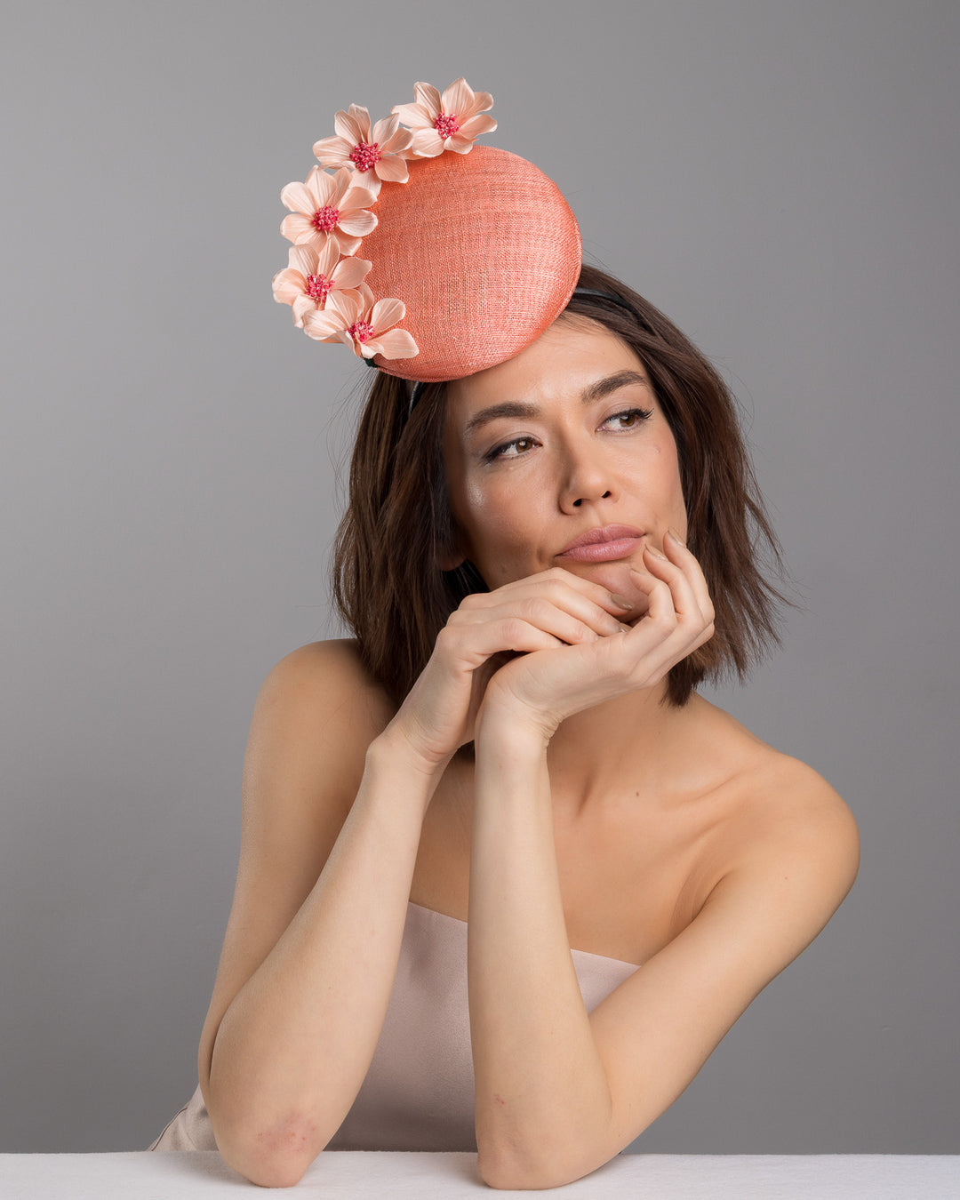 Female model is posing wearing Ainsley coral coloured tall button shaped fascinator attached towards the right side of a headband. The fascinator is decorated from right side to the top with lighter coral coloured five flowers. Designed and made by Merve Bayindir millinery.