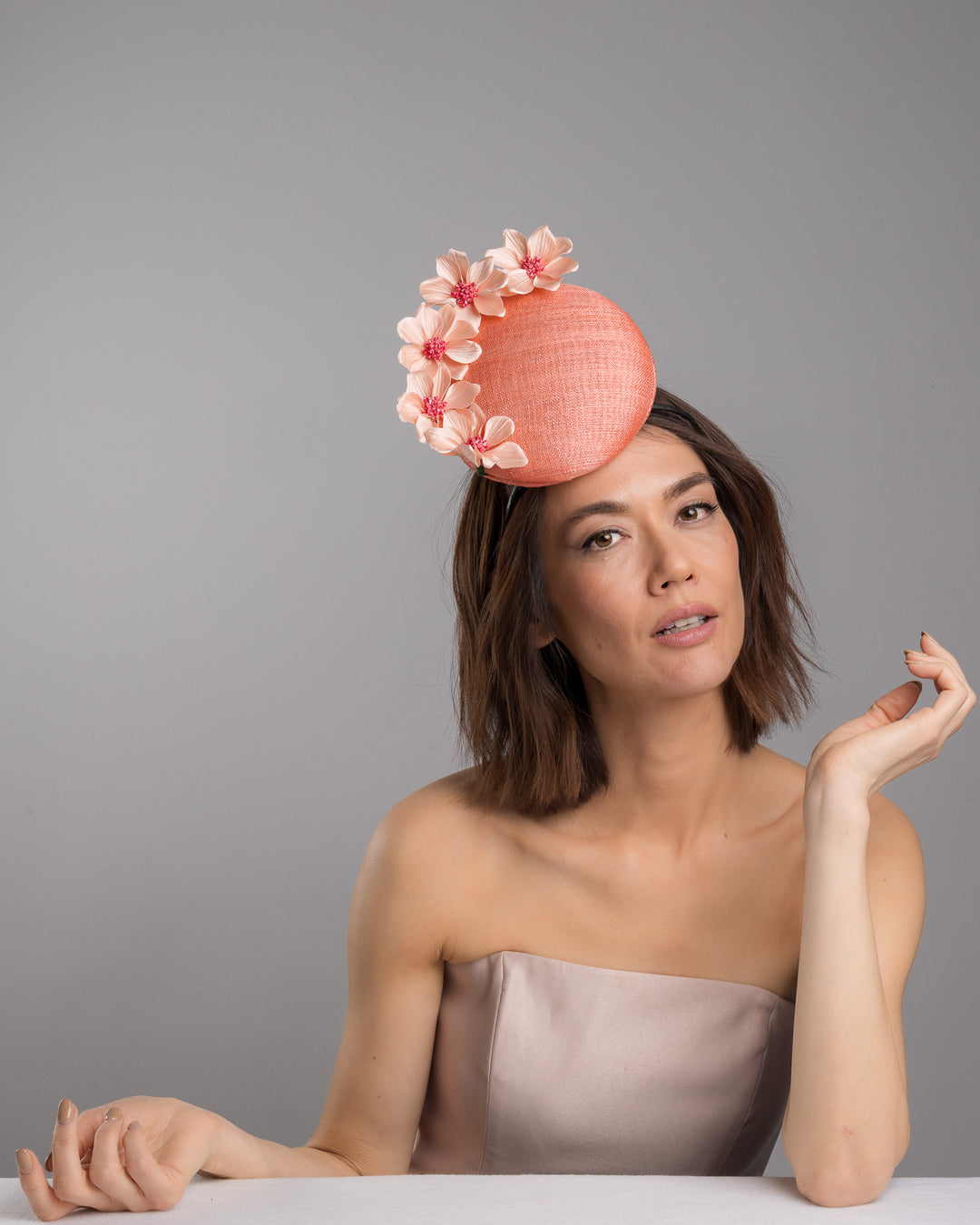 Female model is posing wearing Ainsley coral coloured tall button shaped fascinator attached towards the right side of a headband. The fascinator is decorated from right side to the top with lighter coral coloured five flowers. Designed and made by Merve Bayindir millinery.