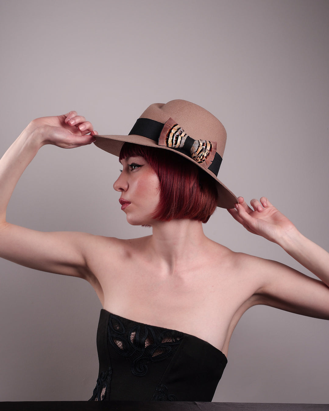 The model is showcasing the right side of the Amelie, a classic fedora hat featuring a feathered bow detail; it&