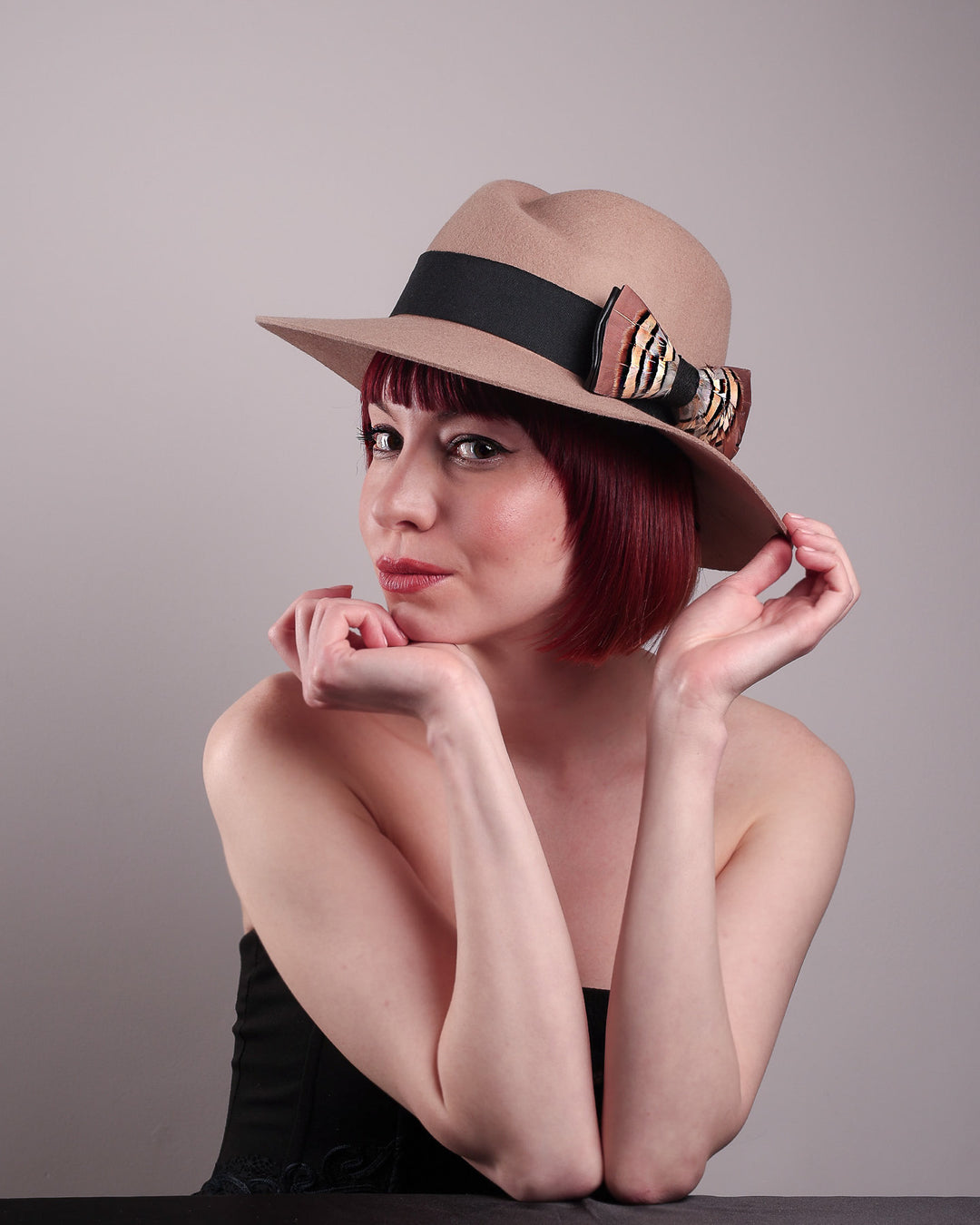 The model is showcasing the left front side of the Amelie, a classic fedora hat featuring a feathered bow detail; it's your go-to when understated charm meets curated flair. Crafted from smooth, light camel felt with a gently pinched crown, this fedora is decorated with a sculptural feather bow, set off against a deep black grosgrain band. Designed by Merve Bayindir.