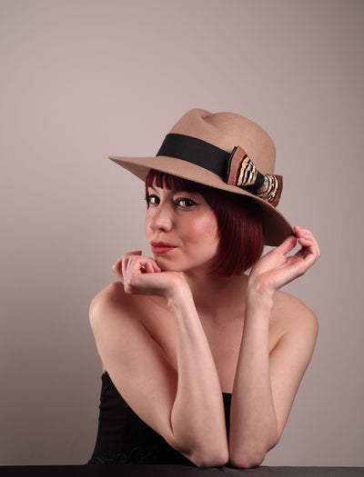 The model is showcasing the left front side of the Amelie, a classic fedora hat featuring a feathered bow detail; it's your go-to when understated charm meets curated flair. Crafted from smooth, light camel felt with a gently pinched crown, this fedora is decorated with a sculptural feather bow, set off against a deep black grosgrain band. Designed by Merve Bayindir.