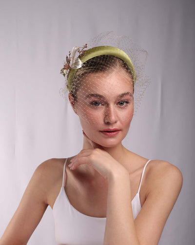 The female model is wearing a sheen green sateen headband decorated with vintage-looking copper-coloured metal leaves and resin flowers on one side. The front and the top of the headband is finished with one-piece sheen green veiling.