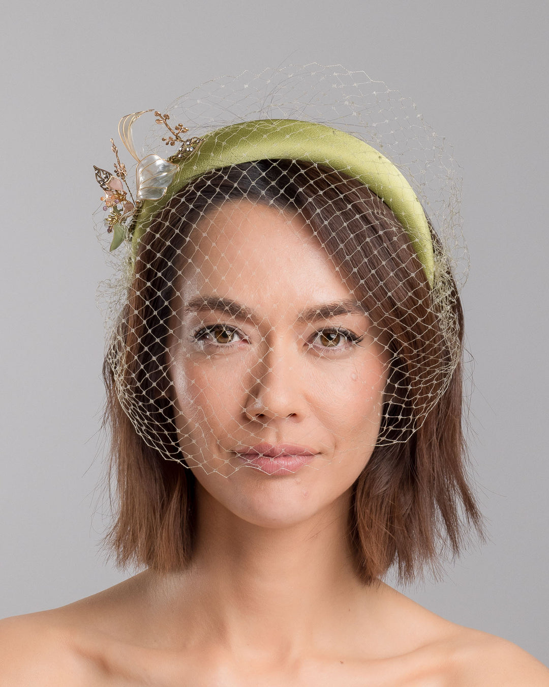 Model is showcasing the front of Amenita Headband , a sheen green silk sateen headband decorated with vintage-looking copper-coloured metal leaves and resin flowers on one side. The front and the top of the headband is finished with sheen green veiling. Made by Merve Bayindir Millinery.