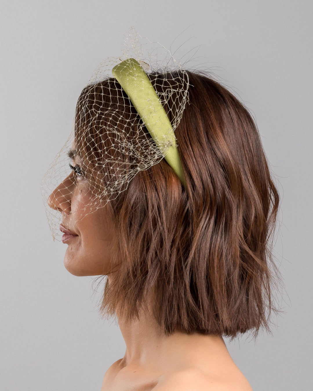 Model is showcasing the left side of Amenita Headband , a sheen green silk sateen headband decorated with vintage-looking copper-coloured metal leaves and resin flowers on one side. The front and the top of the headband is finished with sheen green veiling. Made by Merve Bayindir Millinery.