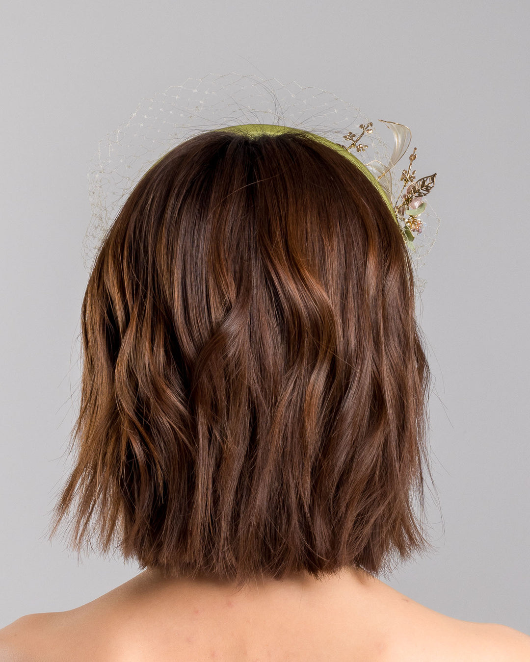 Model is showcasing the back of Amenita Headband , a sheen green silk sateen headband decorated with vintage-looking copper-coloured metal leaves and resin flowers on one side. The front and the top of the headband is finished with sheen green veiling. Made by Merve Bayindir Millinery.