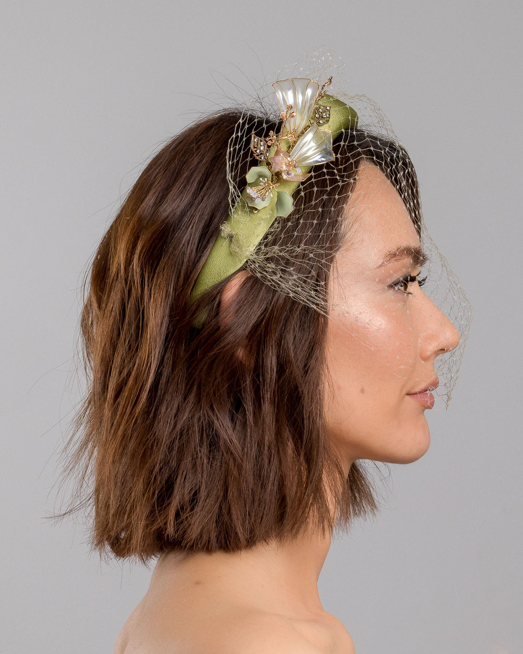 Model is showcasing the right side of Amenita Headband , a sheen green silk sateen headband decorated with vintage-looking copper-coloured metal leaves and resin flowers on one side. The front and the top of the headband is finished with sheen green veiling. Made by Merve Bayindir Millinery.