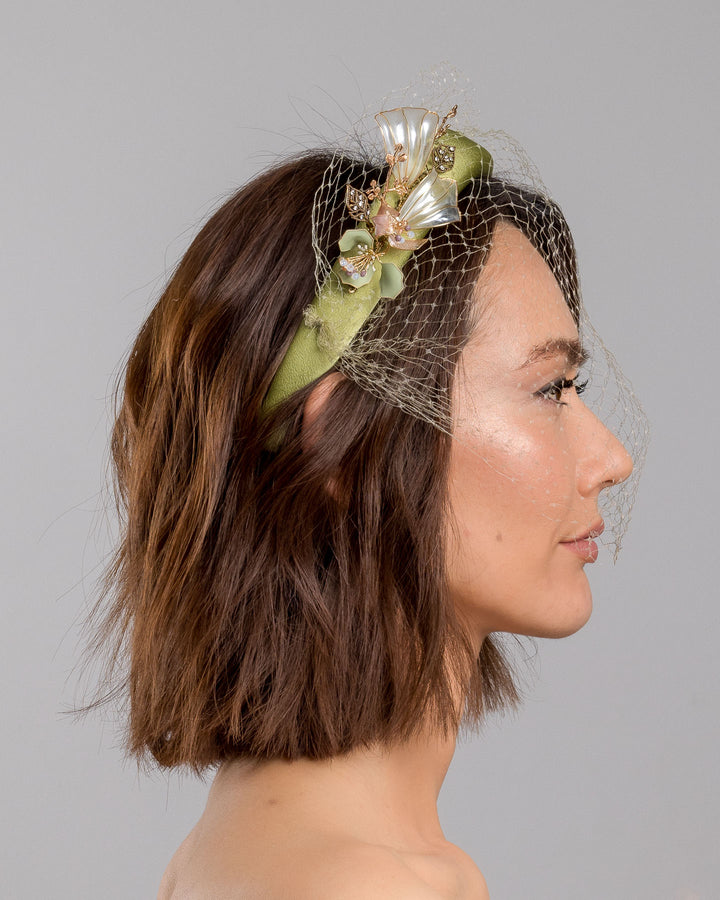 Model is showcasing the right side of Amenita Headband , a sheen green silk sateen headband decorated with vintage-looking copper-coloured metal leaves and resin flowers on one side. The front and the top of the headband is finished with sheen green veiling. Made by Merve Bayindir Millinery.