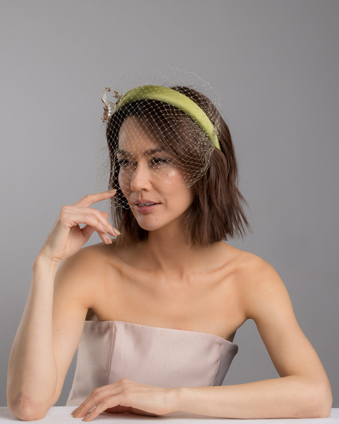 Model is posing wearing Amenita Headband , a sheen green silk sateen headband decorated with vintage-looking copper-coloured metal leaves and resin flowers on one side. The front and the top of the headband is finished with sheen green veiling. Made by Merve Bayindir Millinery.