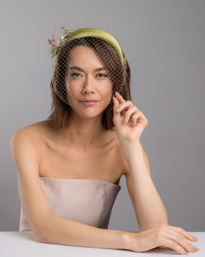 Model is posing wearing Amenita Headband , a sheen green silk sateen headband decorated with vintage-looking copper-coloured metal leaves and resin flowers on one side. The front and the top of the headband is finished with sheen green veiling. Made by Merve Bayindir Millinery.