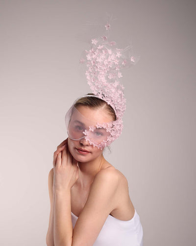  The model is wearing a delicate pink floral mask adorned with tiny flower appliqués extending from the right side of the upper face, cascading upwards and blending seamlessly into a whimsical, semi-transparent veil that gives an ethereal and artistic touch. The mask covers the eyes and nose, enhancing the elegance and beauty of the wearer. The mask is perfect for balls as well as special parties.