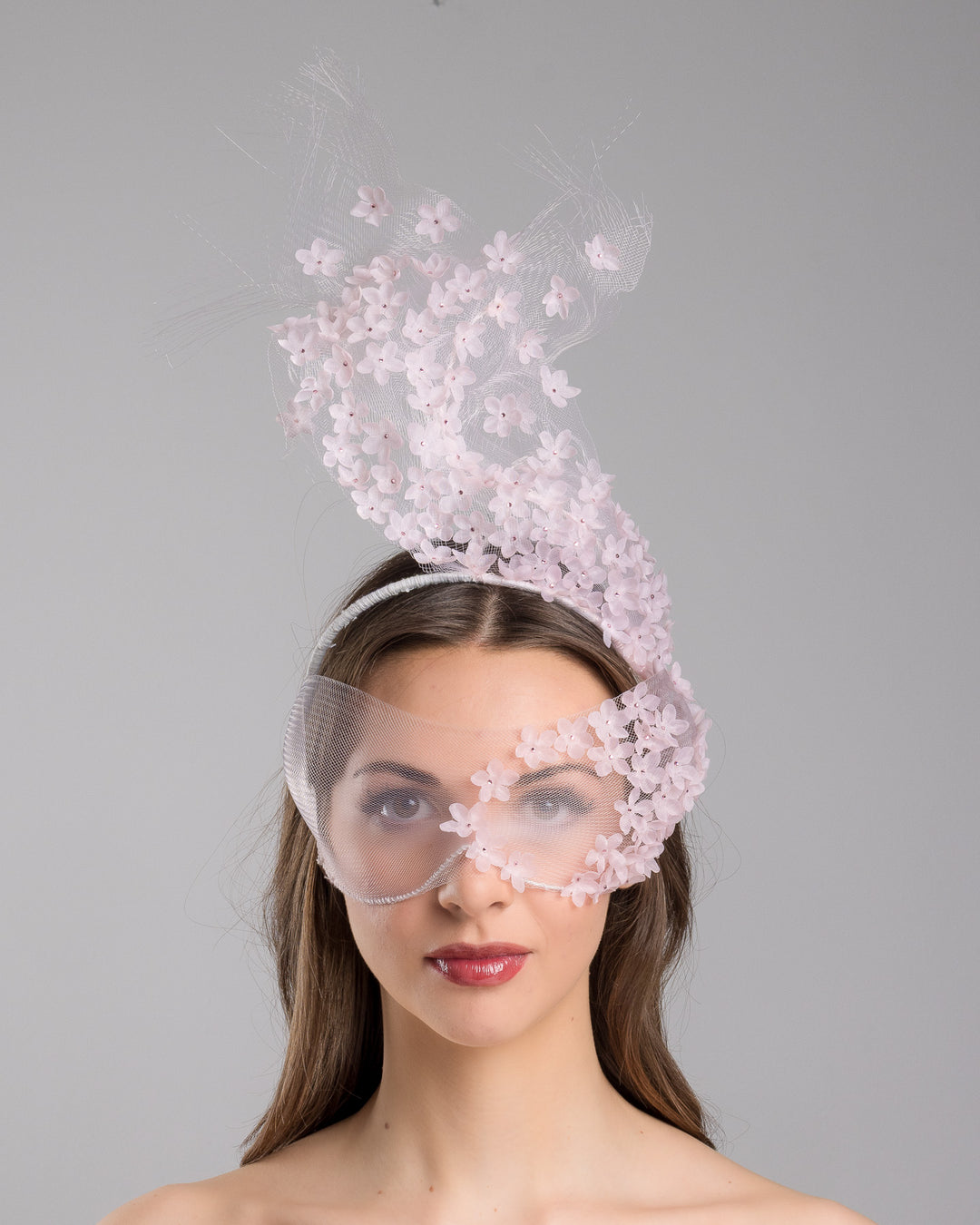 Model is showcasing the front of the delicate couture pink floral sheer mask adorned with tiny flower appliqués with pink crystals extends from the upper face, cascading upwards and blending seamlessly into a whimsical, semi-transparent veil that gives an ethereal and artistic touch. The mask covers the eye part of the face. Made by Merve Bayindir Millinery.
