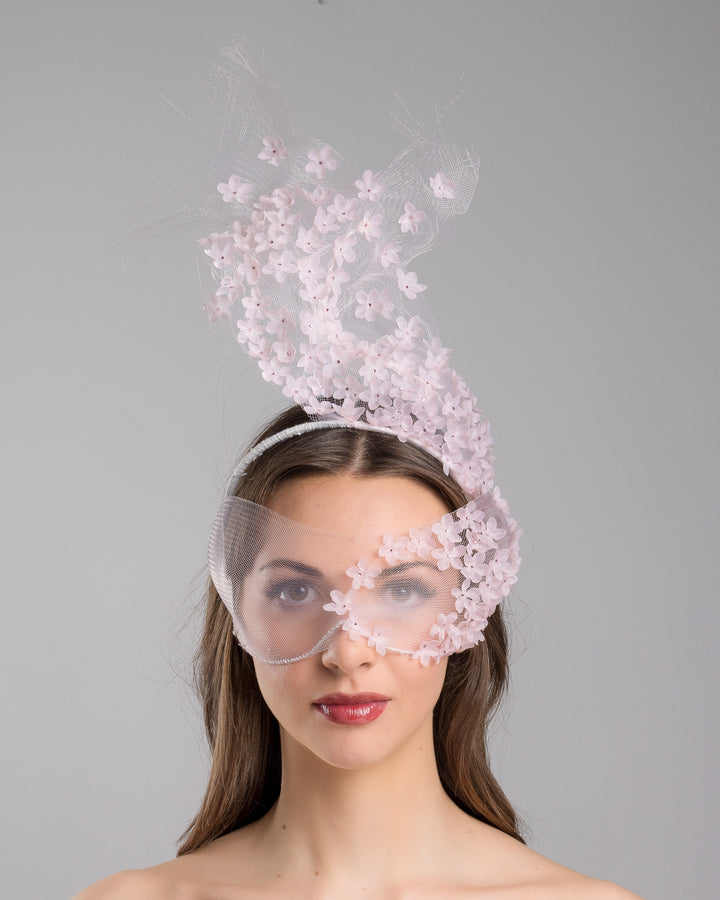Model is showcasing the front of the delicate couture pink floral sheer mask adorned with tiny flower appliqués with pink crystals extends from the upper face, cascading upwards and blending seamlessly into a whimsical, semi-transparent veil that gives an ethereal and artistic touch. The mask covers the eye part of the face. Made by Merve Bayindir Millinery.