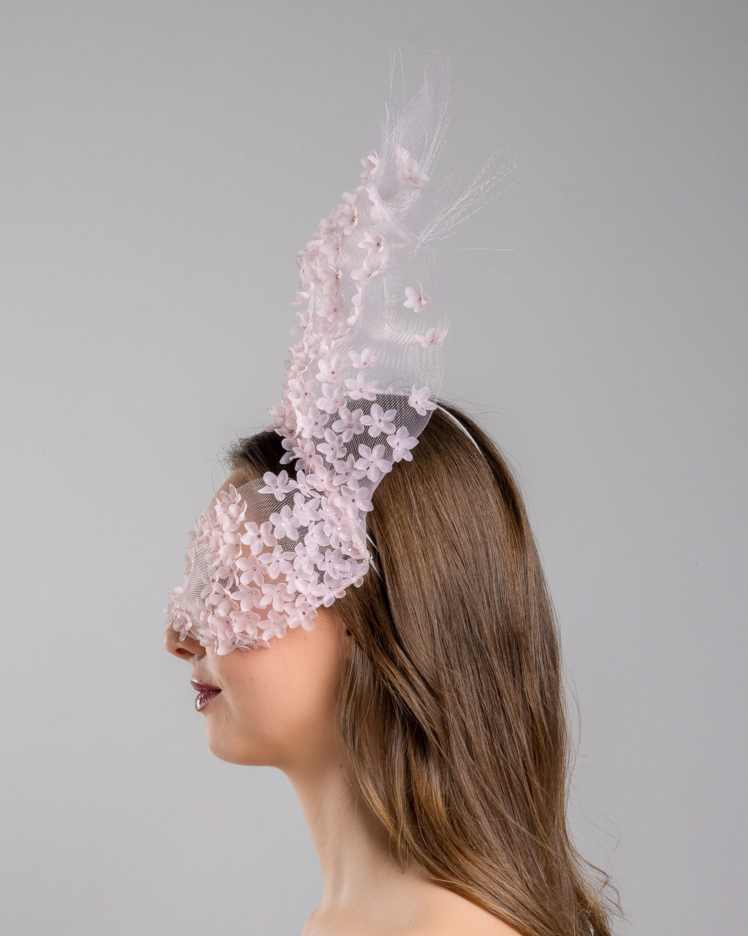 Model is showcasing the left side of the delicate couture pink floral sheer mask adorned with tiny flower appliqués with pink crystals extends from the upper face, cascading upwards and blending seamlessly into a whimsical, semi-transparent veil that gives an ethereal and artistic touch. The mask covers the eye part of the face. Made by Merve Bayindir Millinery.