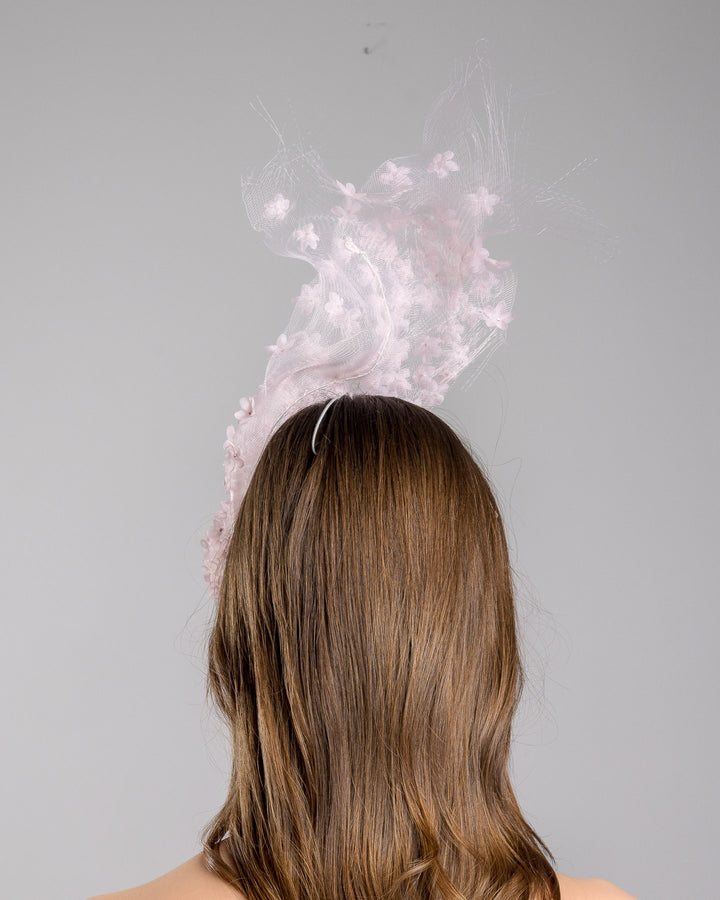 Model is showcasing the back of the delicate couture pink floral sheer mask adorned with tiny flower appliqués with pink crystals extends from the upper face, cascading upwards and blending seamlessly into a whimsical, semi-transparent veil that gives an ethereal and artistic touch. The mask covers the eye part of the face. Made by Merve Bayindir Millinery.
