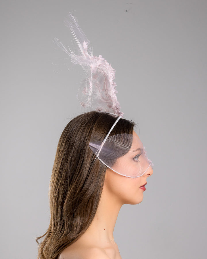 Model is showcasing the right side of the delicate couture pink floral sheer mask adorned with tiny flower appliqués with pink crystals extends from the upper face, cascading upwards and blending seamlessly into a whimsical, semi-transparent veil that gives an ethereal and artistic touch. The mask covers the eye part of the face. Made by Merve Bayindir Millinery.