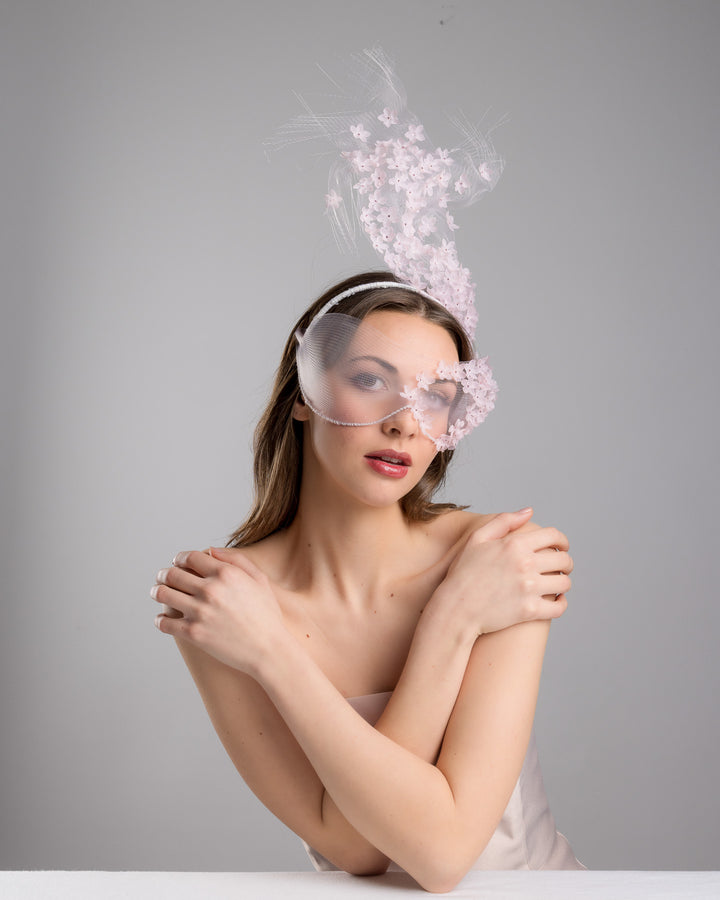 Model is posing wearing  the delicate couture pink floral sheer mask adorned with tiny flower appliqués with pink crystals extends from the upper face, cascading upwards and blending seamlessly into a whimsical, semi-transparent veil that gives an ethereal and artistic touch. The mask covers the eye part of the face. Made by Merve Bayindir Millinery.