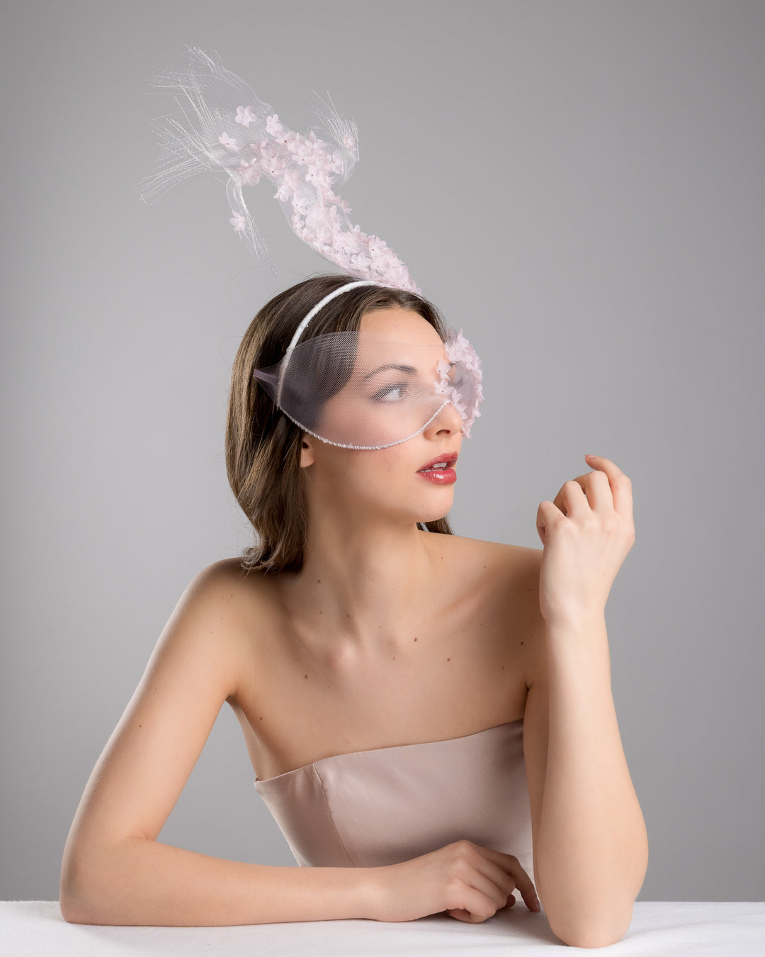 Model is posing wearing  the delicate couture pink floral sheer mask adorned with tiny flower appliqués with pink crystals extends from the upper face, cascading upwards and blending seamlessly into a whimsical, semi-transparent veil that gives an ethereal and artistic touch. The mask covers the eye part of the face. Made by Merve Bayindir Millinery.