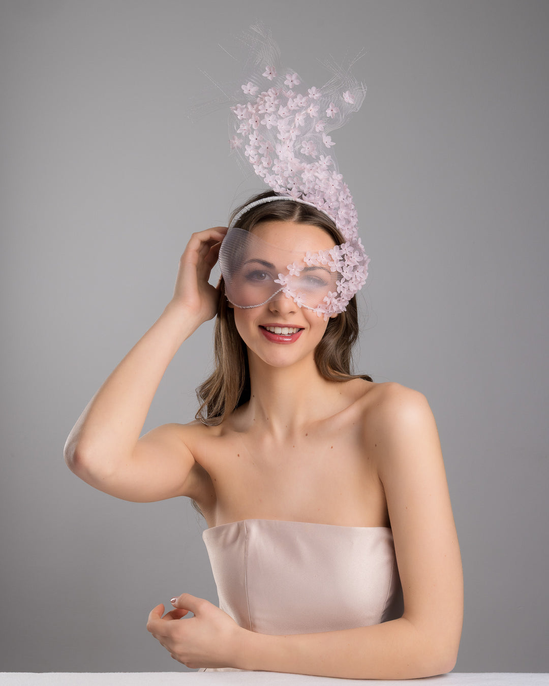 Model is posing wearing  the delicate couture pink floral sheer mask adorned with tiny flower appliqués with pink crystals extends from the upper face, cascading upwards and blending seamlessly into a whimsical, semi-transparent veil that gives an ethereal and artistic touch. The mask covers the eye part of the face. Made by Merve Bayindir Millinery.