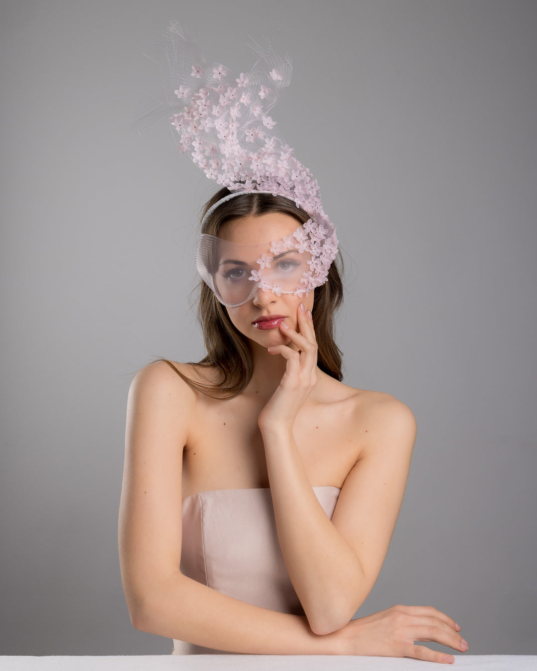 Model is posing wearing  the delicate couture pink floral sheer mask adorned with tiny flower appliqués with pink crystals extends from the upper face, cascading upwards and blending seamlessly into a whimsical, semi-transparent veil that gives an ethereal and artistic touch. The mask covers the eye part of the face. Made by Merve Bayindir Millinery.