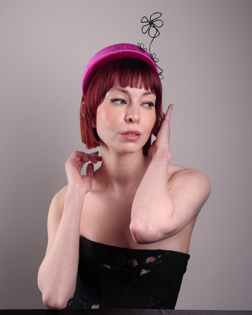 The model, showcasing the right side front of a magenta base, features the Arcana high-formed headband, which sits neatly against the head. At the same time, the matte black floral wire detail ascends upward, accompanied by black wire flowers that create a doodle-like illusion. Designed by Merve Bayindir