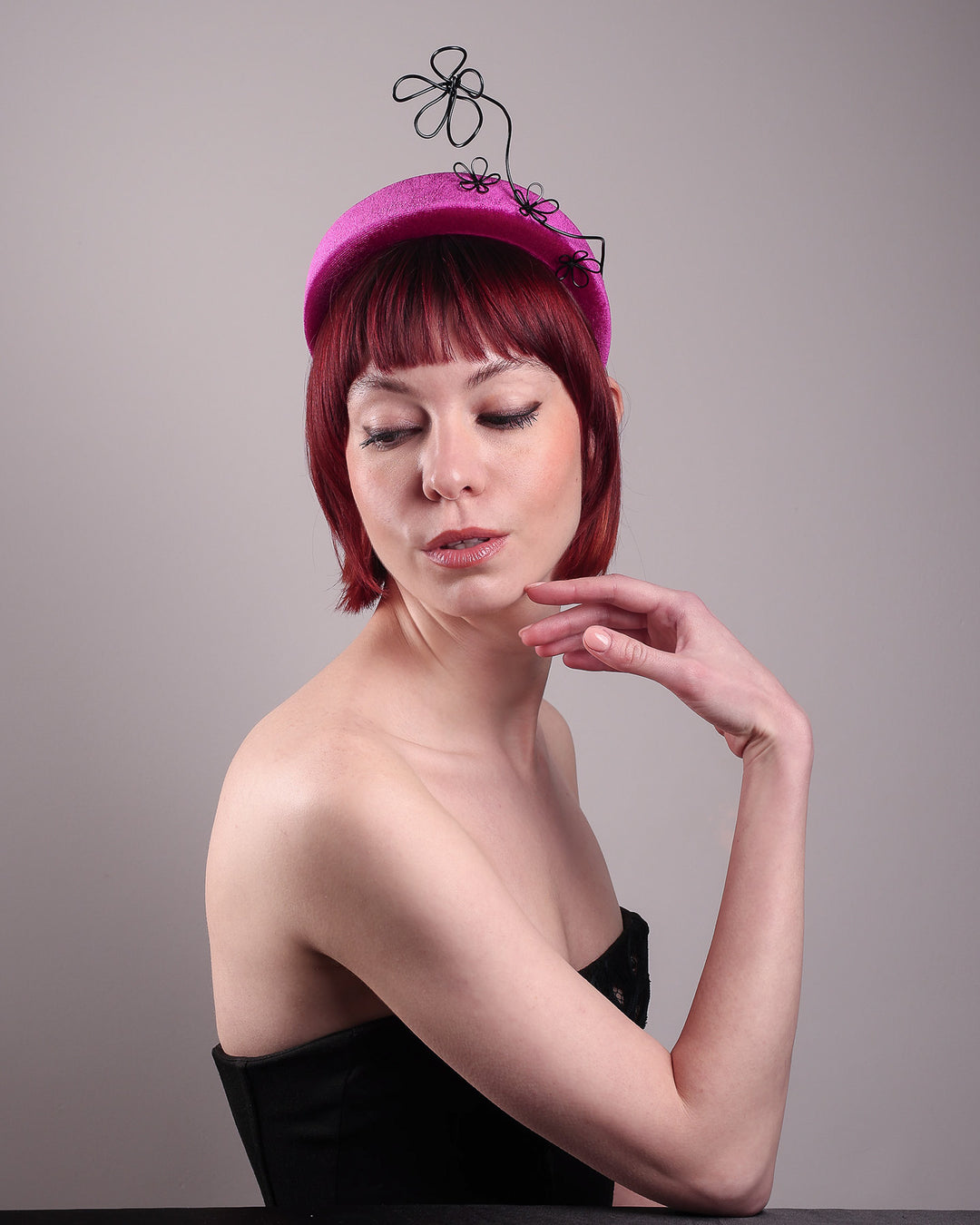 The model, showcasing the left side front of a magenta base, features the Arcana high-formed headband, which sits neatly against the head. At the same time, the matte black floral wire detail ascends upward, accompanied by black wire flowers that create a doodle-like illusion. Designed by Merve Bayindir