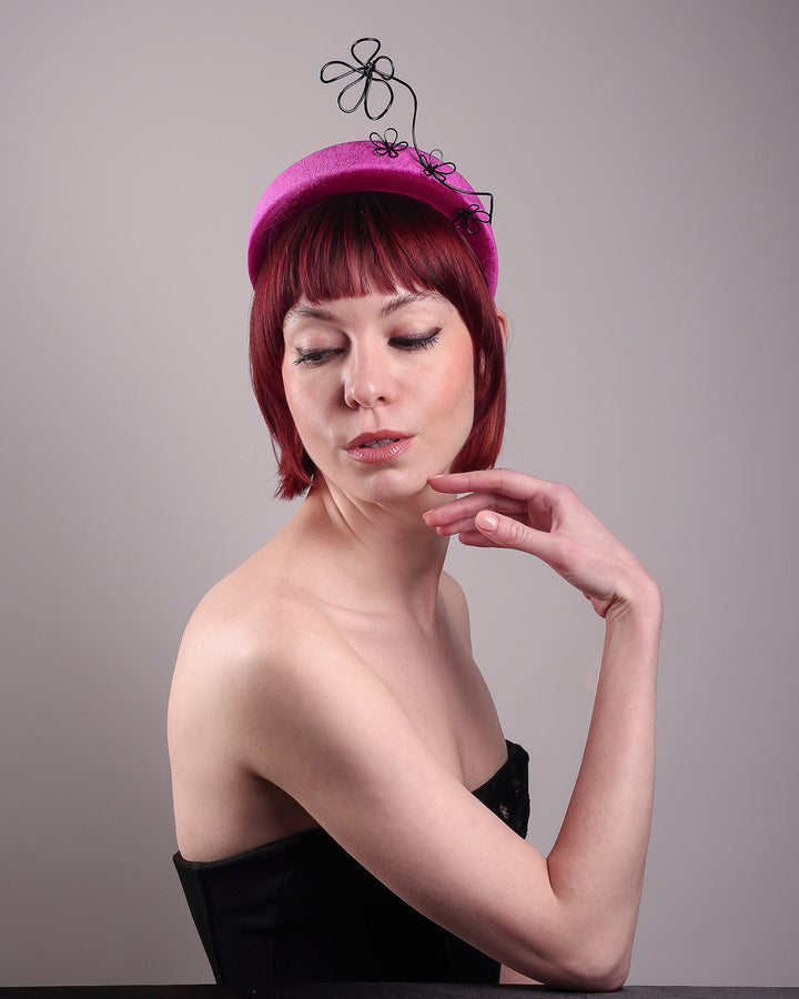The model, showcasing the left side front of a magenta base, features the Arcana high-formed headband, which sits neatly against the head. At the same time, the matte black floral wire detail ascends upward, accompanied by black wire flowers that create a doodle-like illusion. Designed by Merve Bayindir